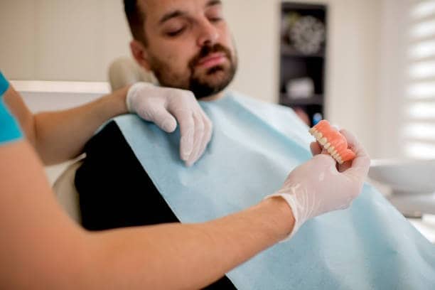 Dentist with gloved hands shows denture model to patient in chair.