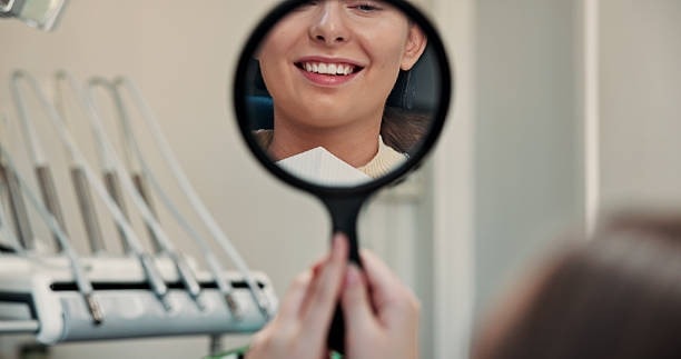 Patient checking smile with handheld mirror in dental chair near instruments