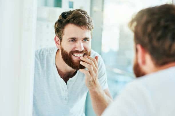 Bearded man smiling at bathroom mirror, checking his teeth with finger