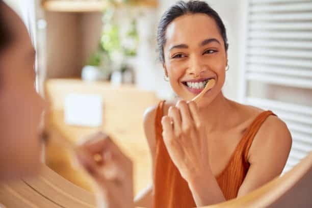 Person brushing teeth with a bamboo toothbrush, smiling at bathroom mirror.