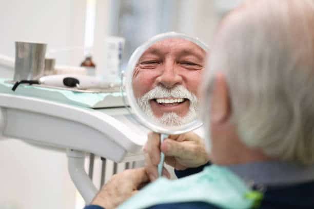 Smiling dental patient examines teeth in mirror in dentist’s chair