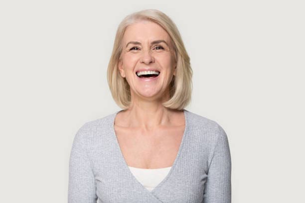 Laughing older woman with gray bob haircut in light sweater, neutral background.