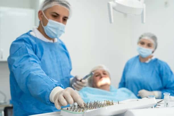 Dentist in mask selects dental implant from sterile kit, patient reclines with assistant nearby.
