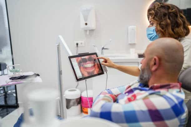 Dentist wearing mask reviews patient’s mouth photo on tablet during consultation.