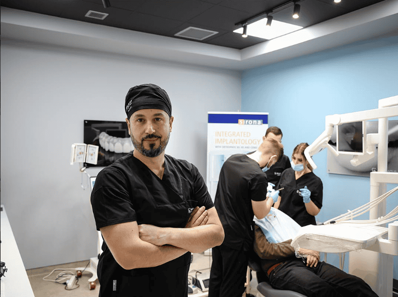 Un dentista con camice nero sta in piedi; il team cura un paziente durante una procedura di impianto dentale.