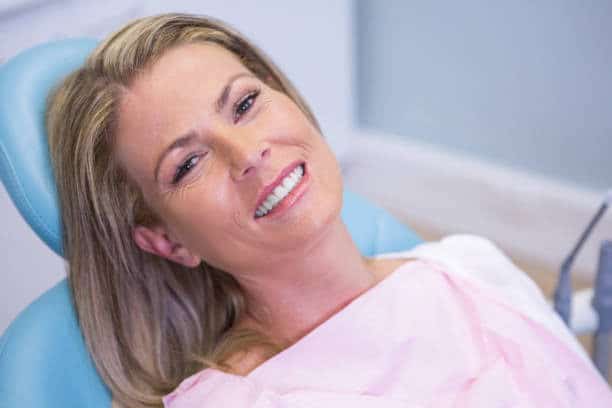 Smiling woman reclining in dental chair, pink bib, bright teeth, modern clinic background.