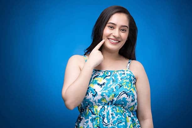 Smiling person in floral dress pointing to healthy teeth against blue background.