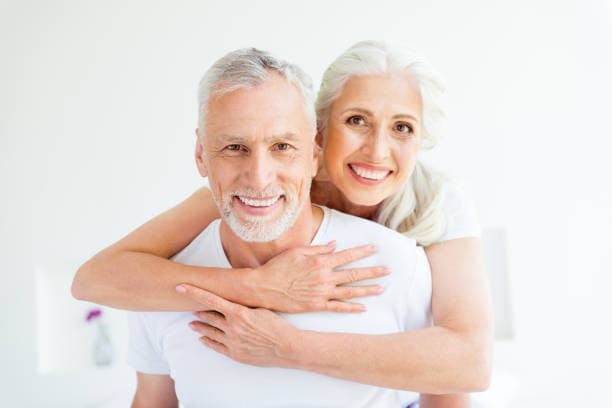 Older couple smiling; woman hugs man from behind in well-lit room.