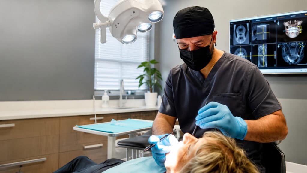Masked dentist treating patient under operatory light, dental implant radiographs and scans on monitor.
