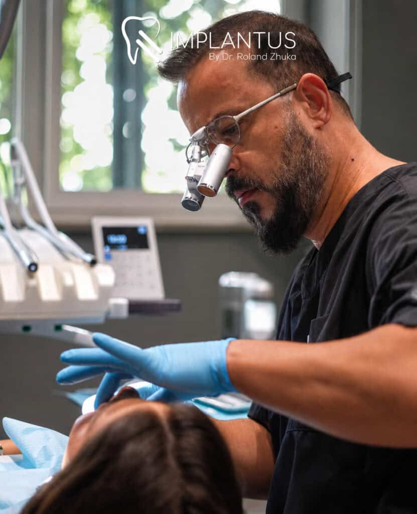 Dentist wearing magnification loupes treats a reclining patient; Implantus Dental Clinic logo visible.