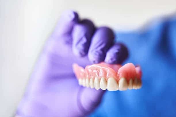 Gloved clinician holding upper full-arch denture, close-up on gum-colored acrylic and teeth.