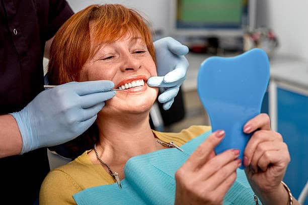 Patient smiling as dentist compares tooth shade guide, holding blue mirror during cosmetic dental check.
