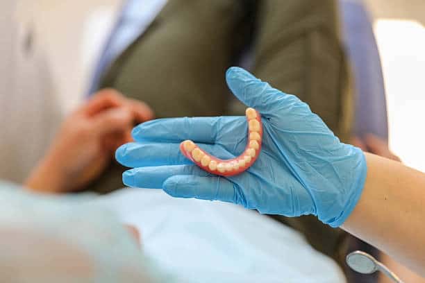 Blue-gloved hand presenting lower implant-supported denture to patient in dental chair.