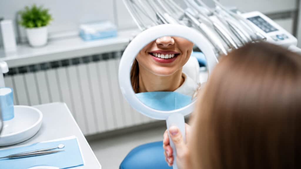 Patient smiling into handheld mirror, wearing bib in dental clinic, instruments on tray.