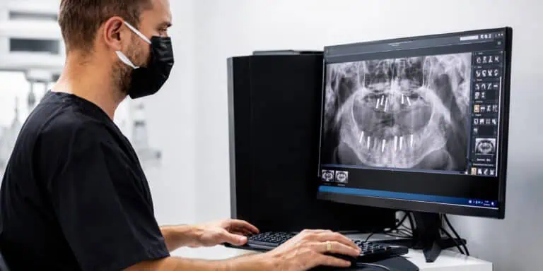 Pacienti admirues i buzëqeshjes së ndritshme në pasqyrë dore në karrigen dentare, me përparëse dhe instrumente.