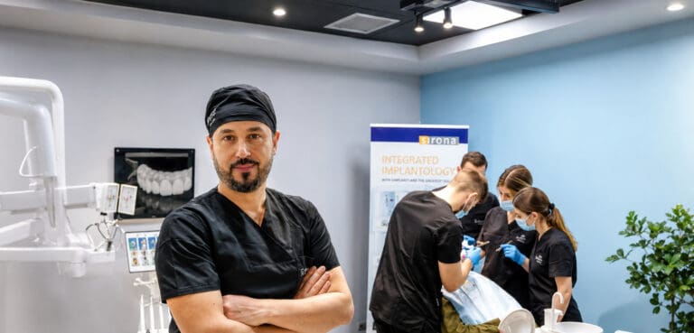 Pacienti duke ekzaminuar buzëqeshjen në një pasqyrë të rrumbullakët të dorës, me përparëse dhe instrumente dentare të dukshme.