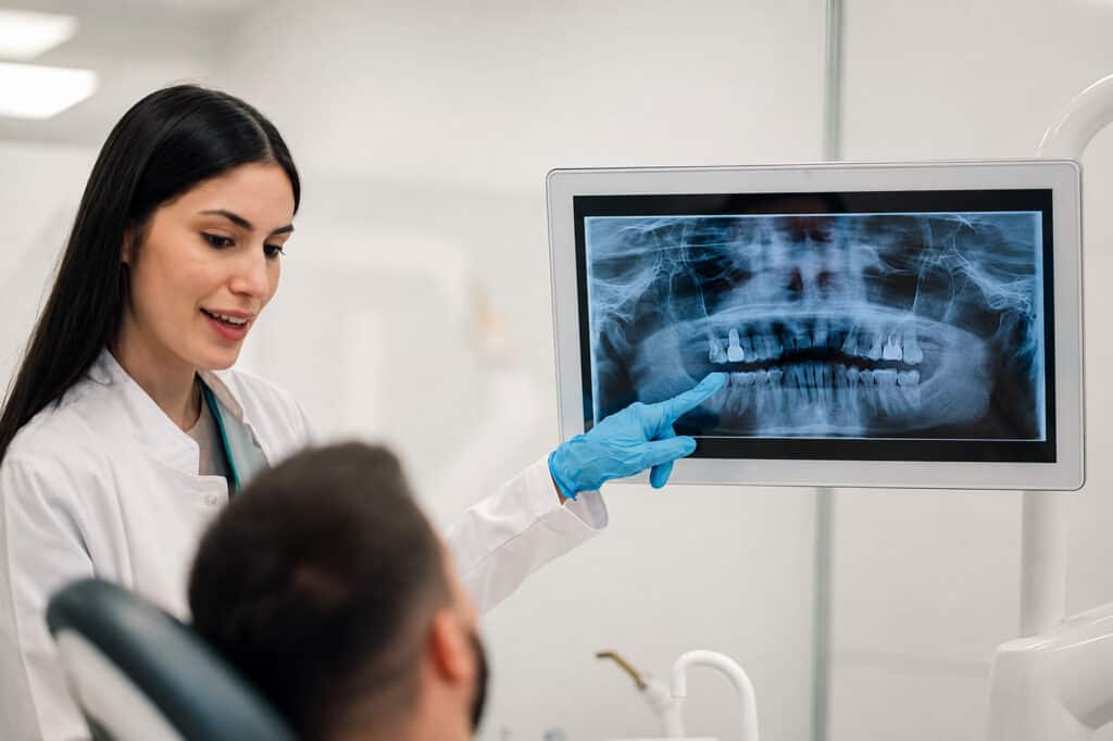 Dentisti me dorezë blu tregon një radiografi panoramike dentare që paraqet implante të shumta.
