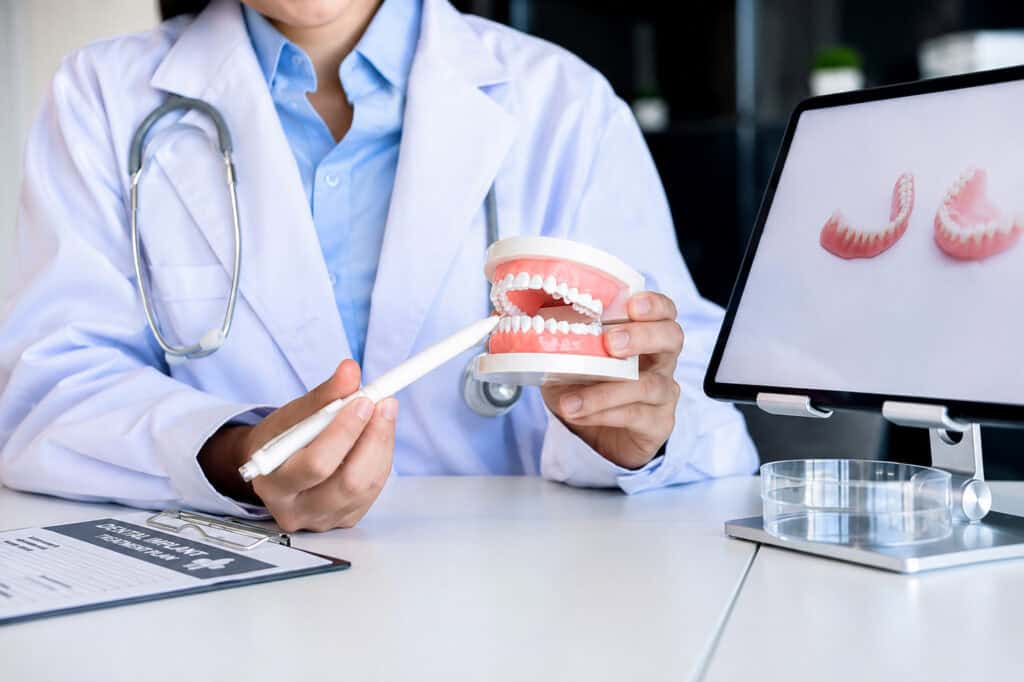 Dentisti shpjegon trajtimin me implante dentare duke përdorur një model të nofullës dhe një tablet që shfaq harqe protezash dentare.