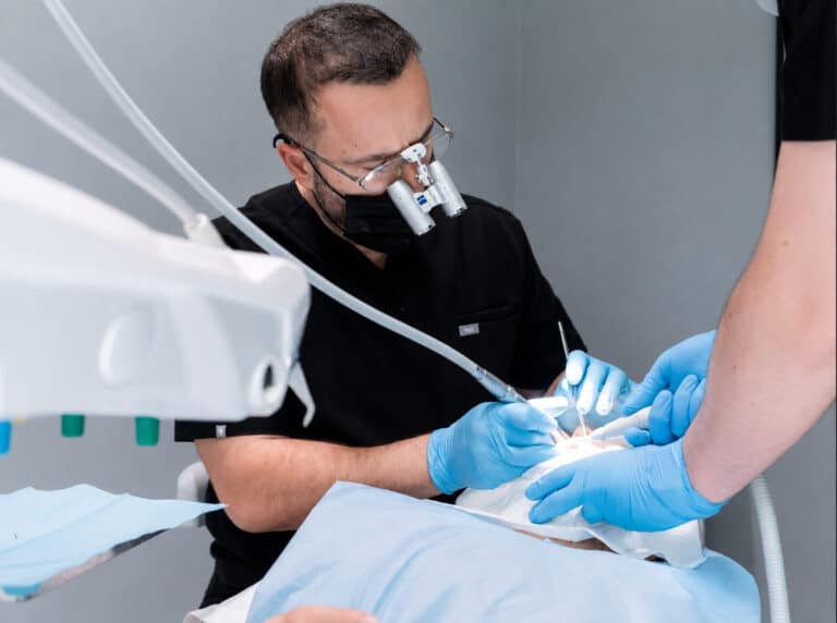Dentisti tregon një model gjatë diskutimit për implantet All-on-4; në monitor shfaqen skanimet e nofullës; pranë karriges ndodhet një valixhe.