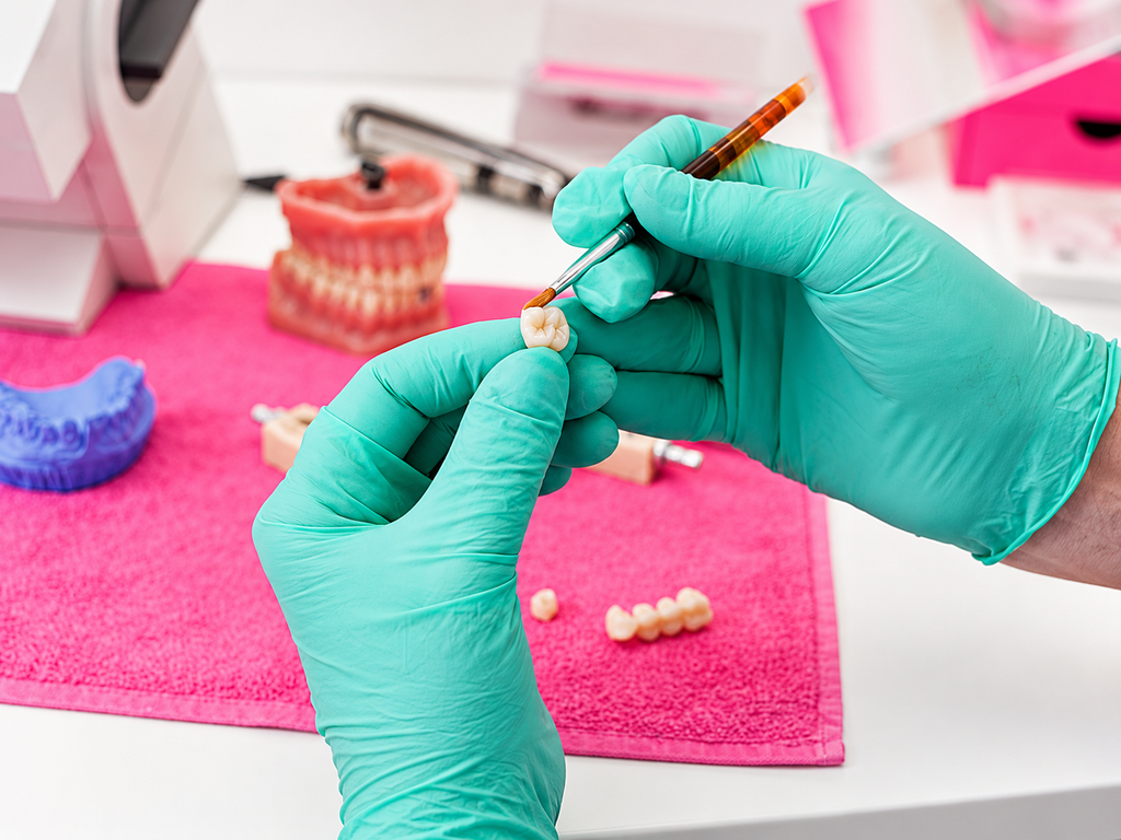 Gloved hands detailing a ceramic dental crown with brush, dental models behind.